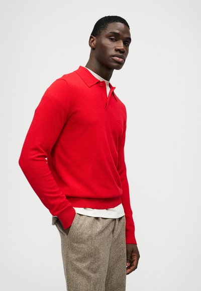 Homem de camisa polo de mangas longas vermelha e calças texturizadas bege, em pé com uma mão no bolso contra um fundo simples.