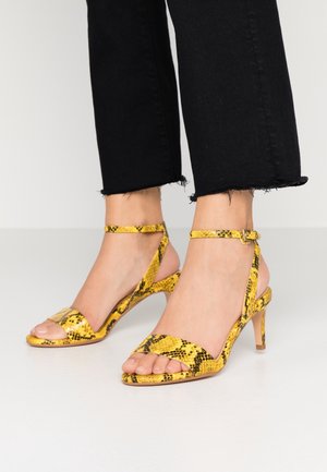 Pieds portant des sandales à talons hauts jaune et noir à imprimé serpent avec brides à la cheville et pantalon noir court à ourlet effiloché.