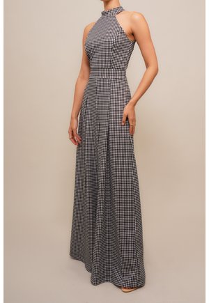 Femme portant une robe maxi sans manches, à col haut, avec un motif pied-de-poule noir et blanc, debout sur un fond neutre.
