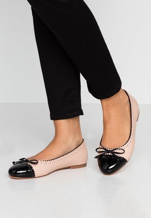 Kvinde iført sorte bukser og tofarvede beige og sorte ballerina sko med sløjfedetaljer, stående på et hvidt gulv.