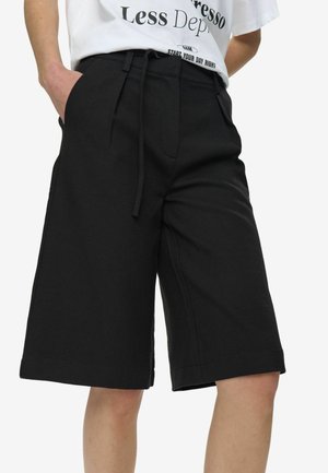 Persoană purtând pantaloni scurți negri largi, până la genunchi, cu talie cu șnur și un tricou alb cu grafică băgat în pantaloni.