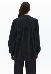 Černá denimová košile s dlouhými, vypouklými rukávy a nařaseným zadním dílem. Má hladkou texturu a volný střih.