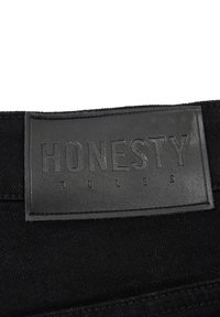 Sort denim talje-label med præget tekst "ÆRLIGHED REGNER" på et mat sort læderpatch, med synlige sømme langs kanterne.