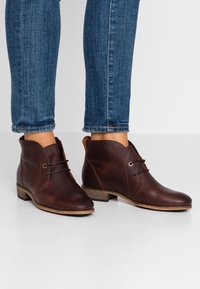 Stivaletti in pelle marrone con punta rotonda, dettagli di allacciatura discreti e tacco in legno basso. Texture liscia e bordo superiore curvato e alla moda.