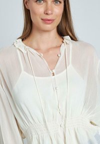 Witte, lichte blouse met een ruches rond de hals, knoopdetails en een gerimpelde taille. Heeft lange mouwen en een zachte, gestructureerde stof.