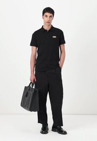 Zwarte poloshirt met een eenzijdig gouden logo op de linkerborst, gecombineerd met losse zwarte broek en een zwarte leren tas.