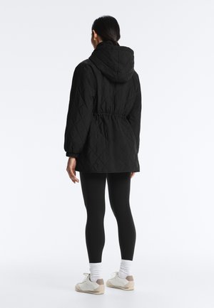 Femeie văzută din spate, purtând o geacă neagră matlasată cu glugă, colanți negri, șosete albe și adidași bej și albi pe un fundal alb.