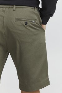 Shorts vert olive en tissu texturé, dotés d'une poche arrière avec un bouton noir et une petite bande métallique accentuée.