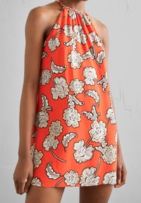 Femme portant une robe sans manches à col licou, orange avec un motif floral blanc et marron, sur un fond uni clair.