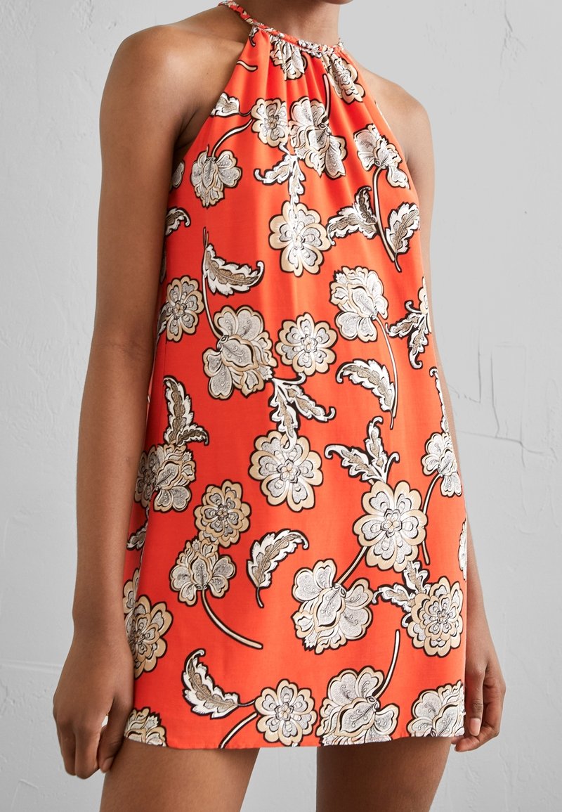 Femme portant une robe sans manches à col licou, orange avec un motif floral blanc et marron, sur un fond uni clair.