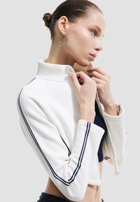 Weißer Zip-Up-Jacke mit hohem Kragen und marineblauen Streifen entlang der Ärmel. Weiches Material mit körperbetontem Design und cropped Länge.