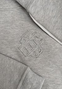 Grijze sweatshirtstof met een gestikte, verhoogde geometrische embleem op de borst en ribgebreide manchetten aan de opgerolde mouwen.