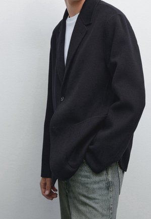 Person trägt einen schwarzen Blazer über einem weißen Hemd und hellblauen Jeans, steht mit einer Hand in der Tasche vor einem einfarbigen Hintergrund.