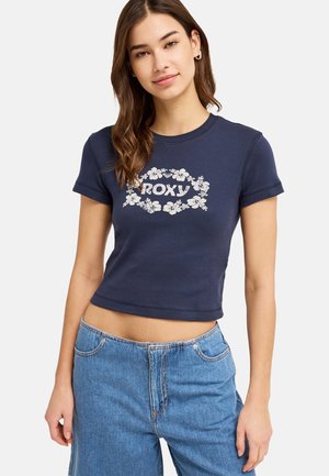 Junge Frau trägt ein marineblaues kurzes T-Shirt mit weißem floralen „ROXY“-Logo und lockere hellblaue Jeans, steht vor einfachem Hintergrund.