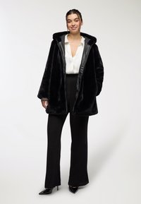 Czarny płaszcz z sztucznego futra z kapturem, z gładką podszewką, noszony na białej bluzce i czarnych spodniach z wysokim stanem, dopełniony czarnymi szpilkami.