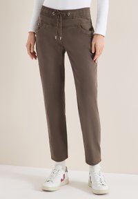 Femme portant un pantalon marron à cordon, une chemise blanche à manches longues, des chaussettes blanches et des baskets blanches avec des accents bordeaux.
