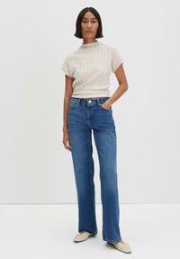 Lichtbeige geribbelde top met korte mouwen, gecombineerd met hoog-gepaste, rechte blauwe spijkerbroek. Model staat casual met de handen in de pockets.