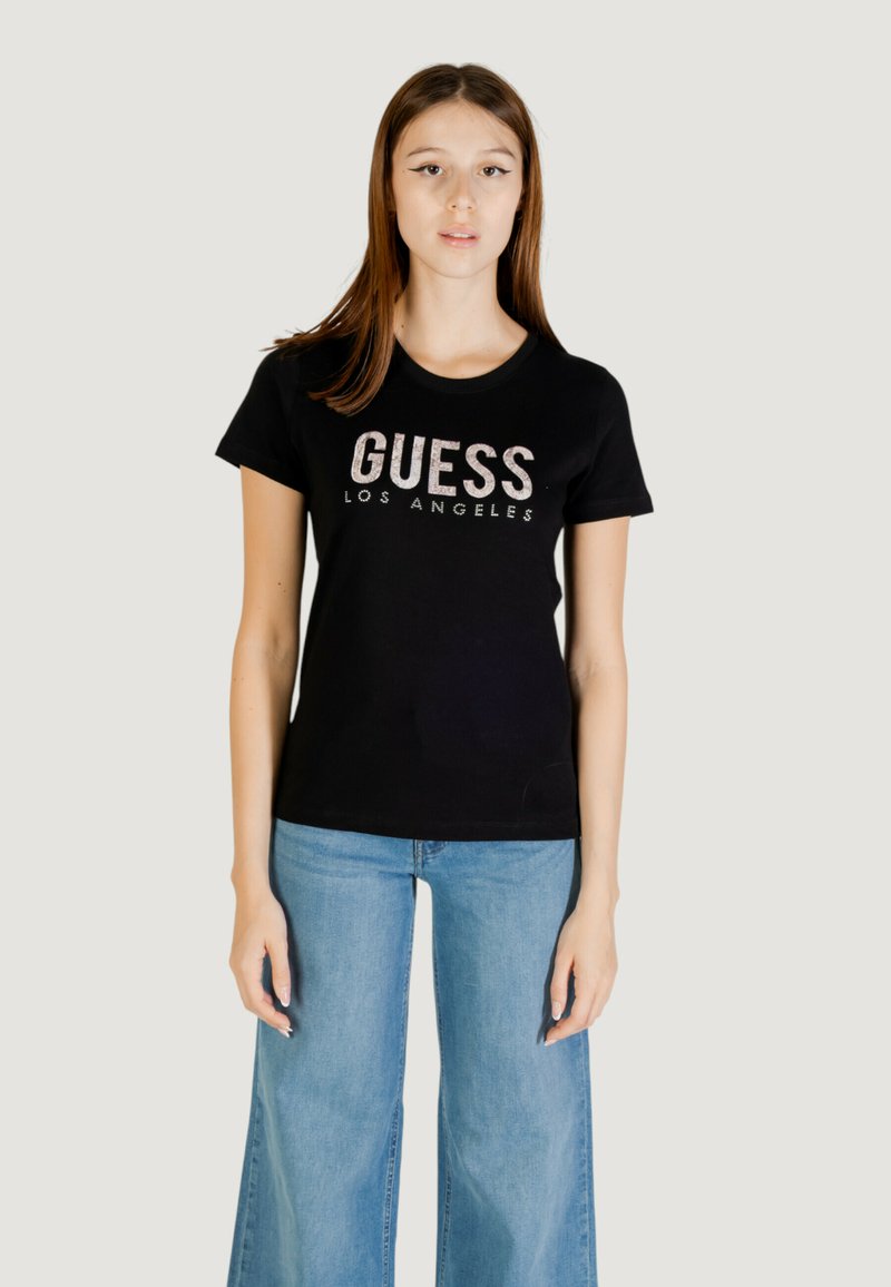 T-shirt in cotone nero con la scritta "GUESS LOS ANGELES" in lettere argento. Il modello indossa jeans a gamba larga blu chiaro. Texture morbida e vestibilità classica.