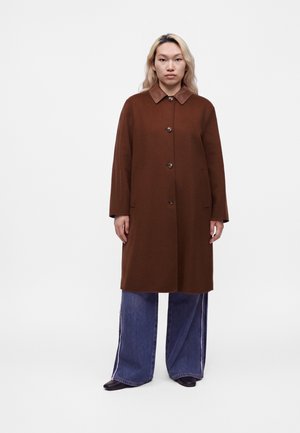 Femme aux cheveux blonds mi-longs, portant un long manteau marron boutonné, un jean large bleu et des chaussures noires, debout devant un fond uni.