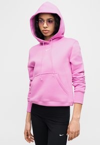 Rosa hettegenser laget av mykt stoff med en frontlomme, snorer og elastiske mansjetter; brukt sammen med svarte leggings med en logo.