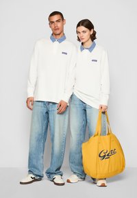 Camisas de polo de manga larga en blanco con cuellos en azul claro, combinadas con jeans azul claro. Bolsa de mano amarilla con el texto náutico "Gant". Calzado: zapatillas blancas.