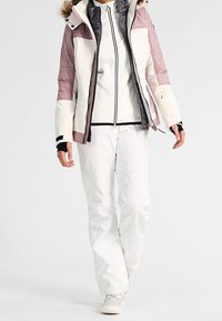 Veste matelassée blanche et rose clair avec capuche bordée de fourrure, design matelassé, sections sombres contrastantes. Pantalon et chaussures blancs, silhouette élégante.