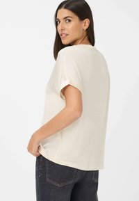 Cream-colored T-shirt with rolled cuffs, relaxed fit, soft texture, and a slightly longer back hem, paired with dark denim jeans.