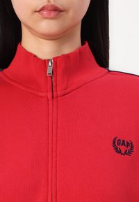 Giacca rossa con zip in cotone, dotata di colletto alto, con zip argentata e dettagli blu navy sulle spalle; logo GAP ricamato in blu navy.