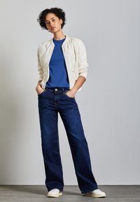 Světle krémová prošívaná bomber bunda přes modré tričko, doplněná tmavě modrými širokými džíny a bílými teniskami. Jednoduchý a moderní design.