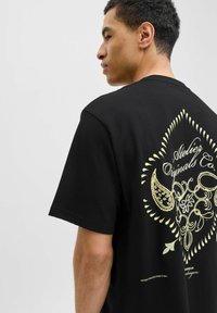 Sort bomulds t-shirt med et stort, intrikat gult grafisk design på ryggen. Korte ærmer og en afslappet pasform.