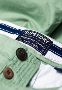 Lichtgroene chino broek met een marineblauw label. Voorzien van een donkerbruine knoop en witte stikaccenten. Het label draagt de tekst "Superdry Premium Chino, New York."