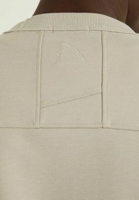 Felpa beige con scollatura a girocollo a coste. Dettaglio posteriore notevole con un patch triangolare cucito e leggeri accenti di ricamo.