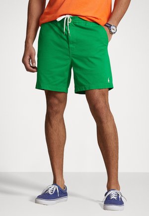 Man draagt felgroene shorts met wit trekkoord, oranje shirt, blauwe sneakers en polshorloge, staand tegen een witte achtergrond.