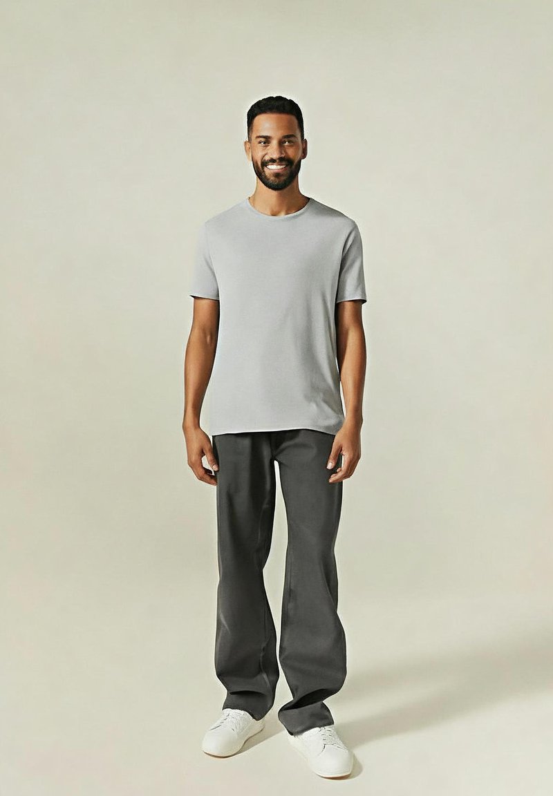 Homme souriant debout, portant un t-shirt à manches courtes gris clair, un pantalon ample sombre et des baskets blanches sur un fond neutre.