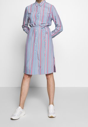 Personne portant une robe-chemise à manches longues rayée verticalement bleu, rose et blanc avec des boutons et des baskets blanches, debout les mains dans le dos.