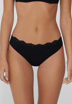 Black lace-trimmed bikini underwear worn by a woman, highlighting the midsection and upper thighs against a plain background.