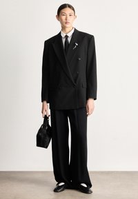 Μαύρο σακάκι με διπλό πέτο και φαρδιές πέτρες, συνδυασμένο με χαλαρά μαύρα παντελόνια. Λευκό πουκάμισο και γραβάτα από κάτω, κρατώντας μια μικρή μαύρη τσάντα.