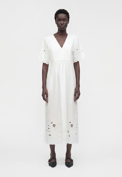 Femme portant une robe midi blanche avec des bords festonnés, des détails floraux en œillets, des manches courtes et des chaussures noires pointues, debout devant un fond uni.