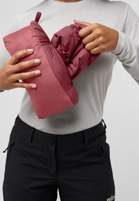 Une veste isolante bordeaux étant emballée en une forme compacte, montrée avec un haut à manches longues gris clair et un pantalon noir. Présente une texture lisse.