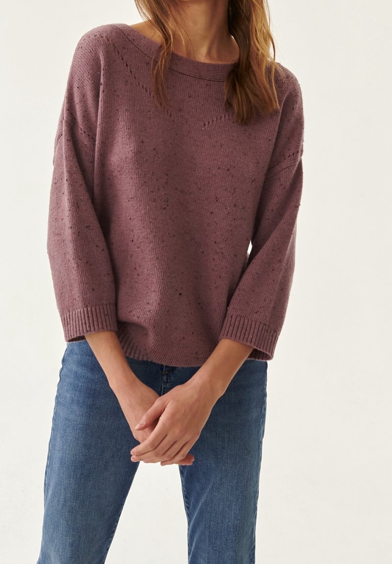 Purple knitted sweater with a relaxed fit, batwing sleeves, and ribbed cuffs. Subtle flecks and detailed texture. Worn with blue denim jeans.