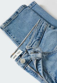 Jean en denim bleu clair plié, avec une fermeture éclair partiellement ouverte, un bouton visible et une étiquette de marque blanche près de la ceinture.