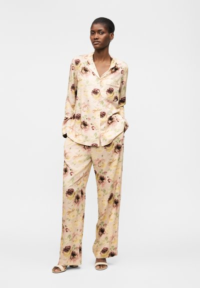 Naine kannab lahtist, lillelist kreemikat siidist pidžaamakomplekti pikkade varrukatega ja laia säärega pükstega, seisab käed taskutes.