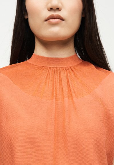 Femme aux longs cheveux foncés portant un haut orange texturé à col montant, visible des épaules jusqu'aux lèvres, sur un fond uni.