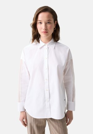 Femme aux cheveux bruns mi-longs porte une chemise blanche boutonnée avec des manches transparentes rayées et un pantalon beige à fines rayures.