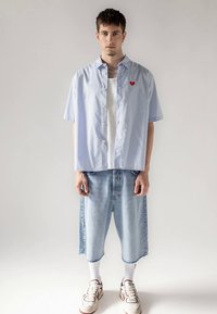 Světle modrá košile s krátkým rukávem na zapínání na knoflíky s červeným srdíčkovým emblémem, doplněná volnými světle modrými džínovými šortkami a bílými ponožkami, zakončená teniskami.