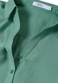 Chemise verte en tissu lisse, dotée d'un col classique, d'une fermeture à boutons et d'une petite poche poitrine. L'étiquette indique la marque "CECIL" et la taille "S."