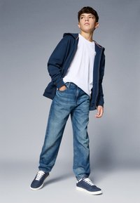 Jachetă cu glugă de culoare bleumarin peste un tricou alb, asortată cu blugi de denim de culoare albastră deschisă și teniși bleumarin cu talpă albă. Stil casual.