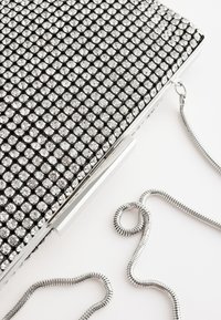 Zilveren clutch tas bedekt met rijen kleine heldere strasstenen met een zilveren slangkettingband op een witte achtergrond.