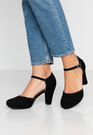 Chaussures à talons hauts en cuir suédé noir avec des brides de cheville, bout arrondi et finition décorative. Associées à un jean court bleu clair.