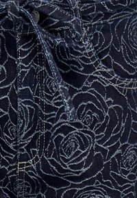 Jeans hlače temno modre barve z belim vzorcem vrtnice, vidnim šivom in detajlom zavezovanja na pasu.